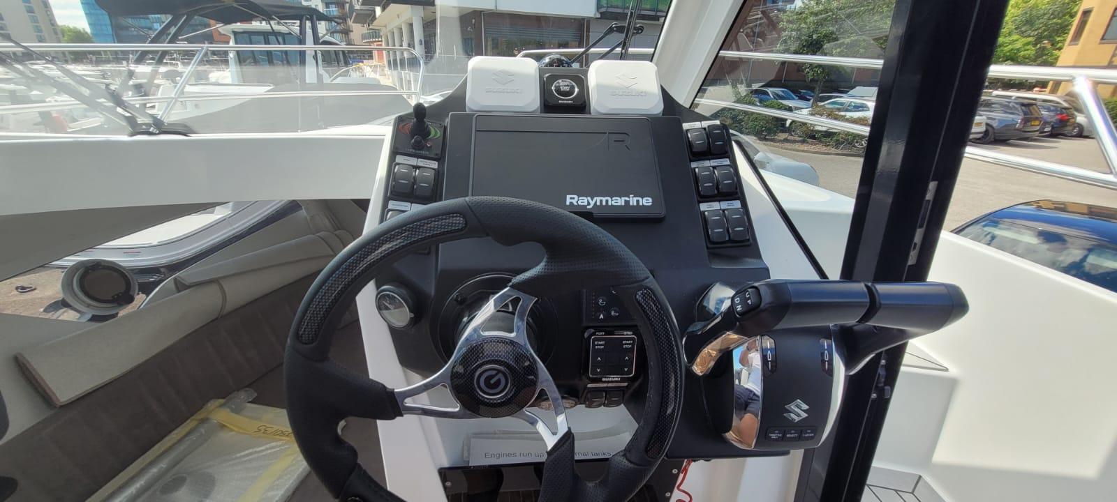 Boat cockpit featuring a Raymarine display, steering wheel, and control levers, with visible logos and adjacent boats.